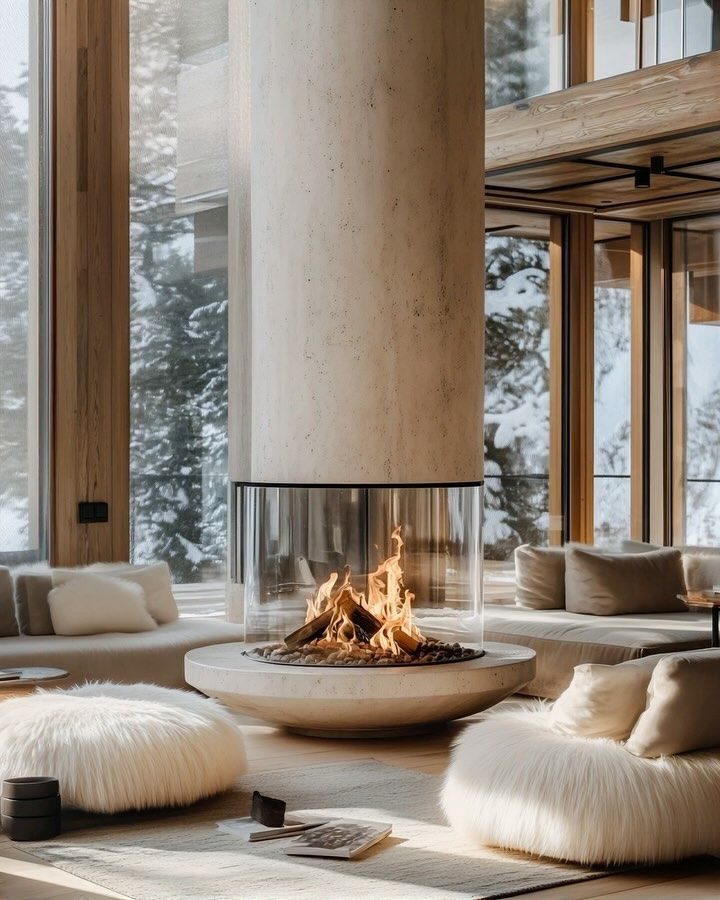 A cylindrical travertine gas fireplace burning behind curved glass in a snowbound chalet.